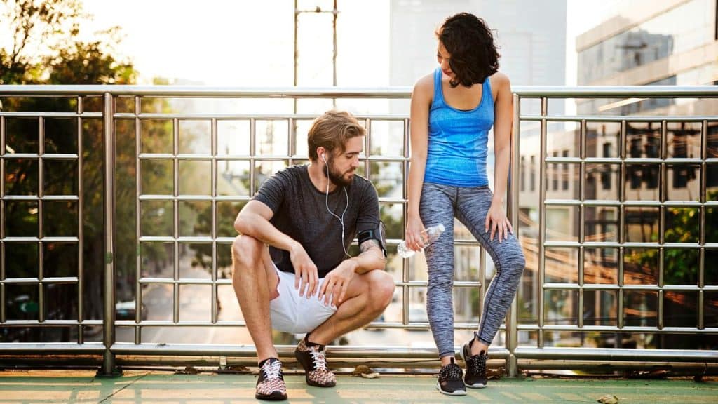 A man crouches and a woman stands together on a bridge in athletic wear.
