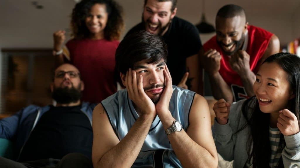 A man rests his head in his hands while several people behind him cheer and celebrate.