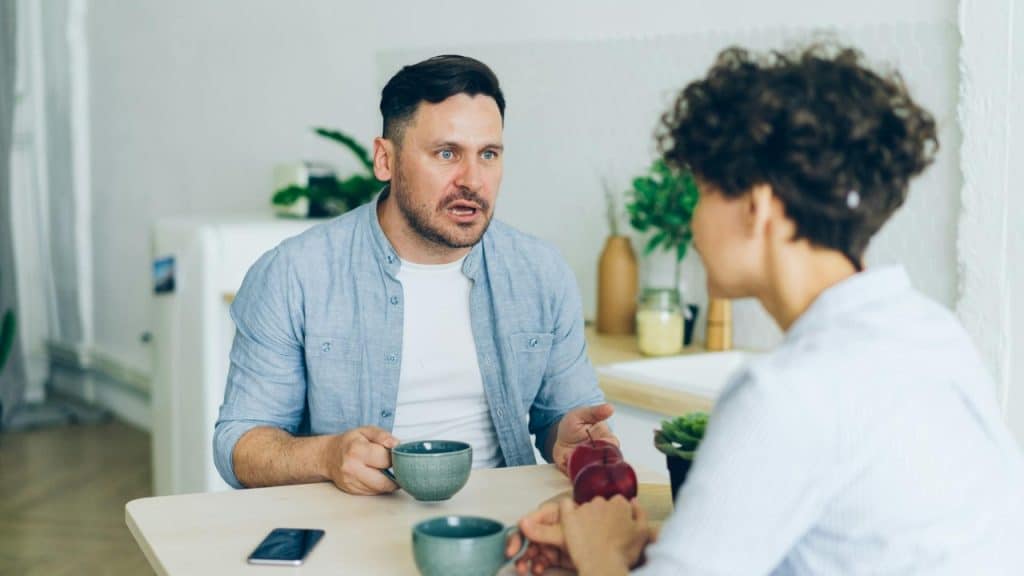 A man speaking expressively while sitting at a table across from a person with curly hair.