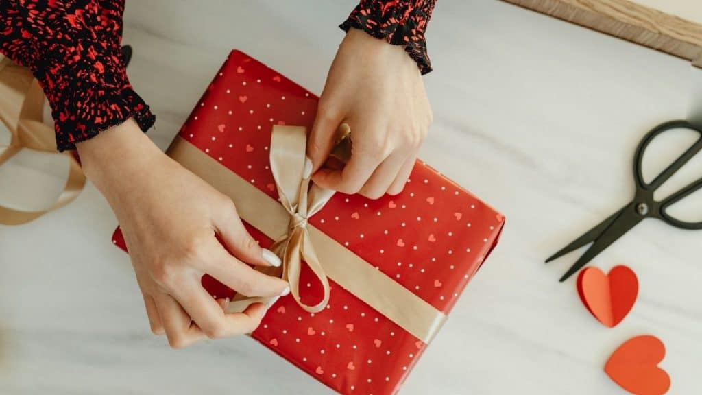 Hands tie a gold ribbon on a red gift box with white heart patterns.