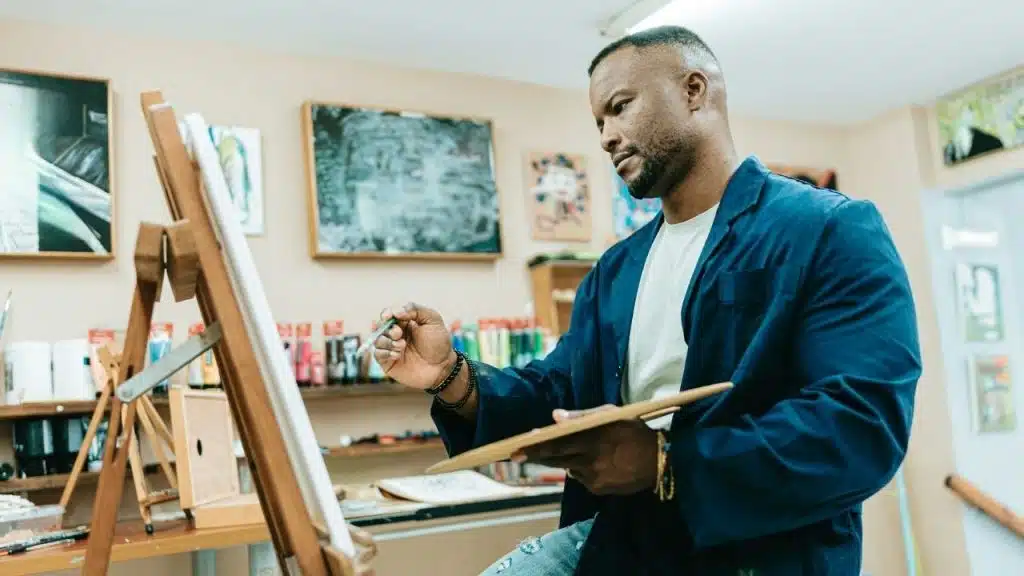 A man in a blue jacket paints on a canvas while holding a wooden palette.