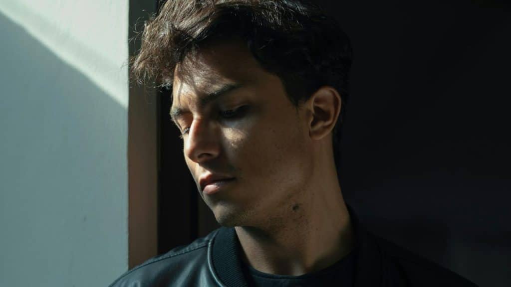 A man in a black jacket looks downward while standing in a beam of sunlight.
