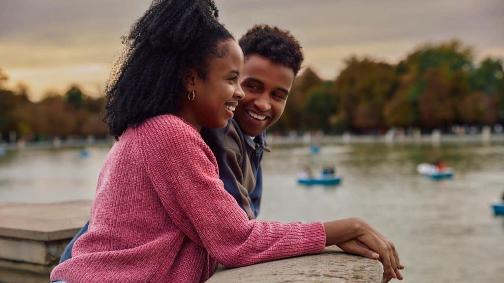 A smiling couple leaning on a railing by a lake at sunset.