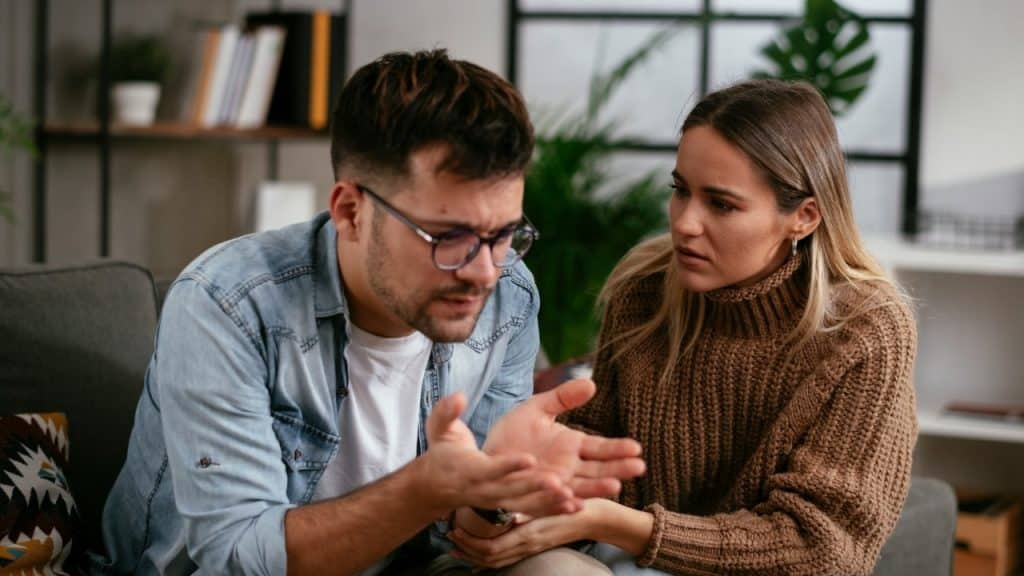 A man with glasses gestures with his hands while sitting on a sofa next to a woman.