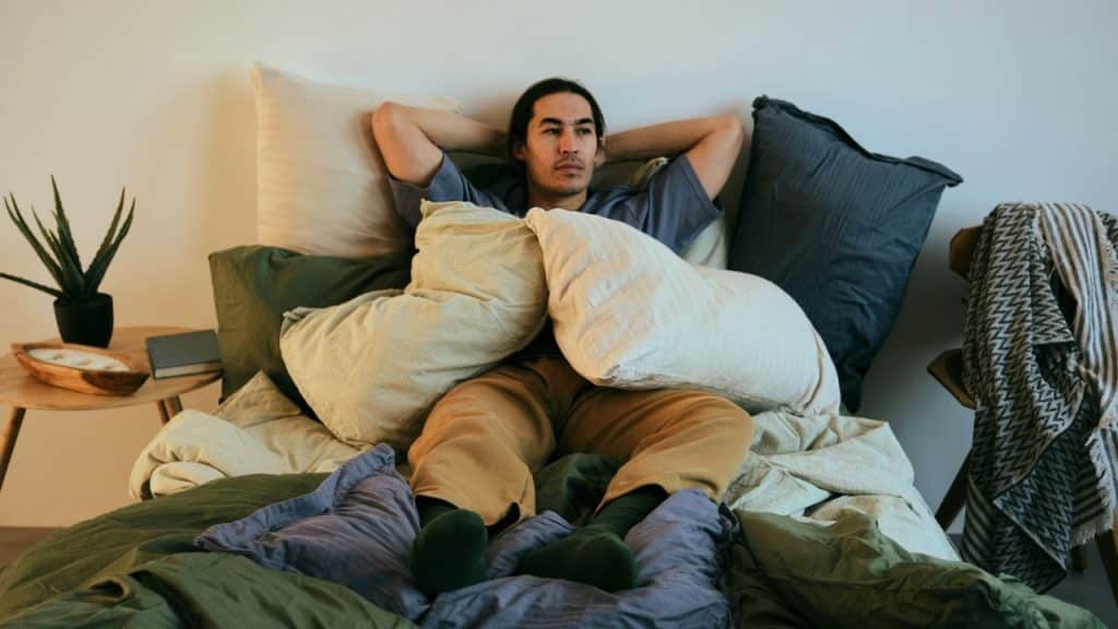 A man reclines on a bed with his hands behind his head, surrounded by pillows.