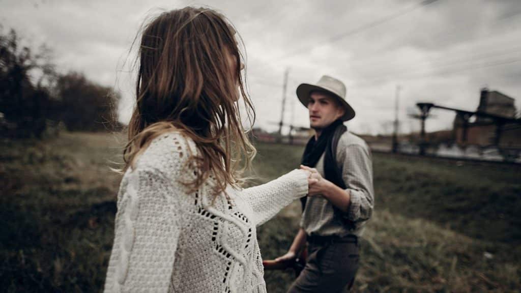 A woman in a white sweater holds hands with a man wearing a hat outdoors.