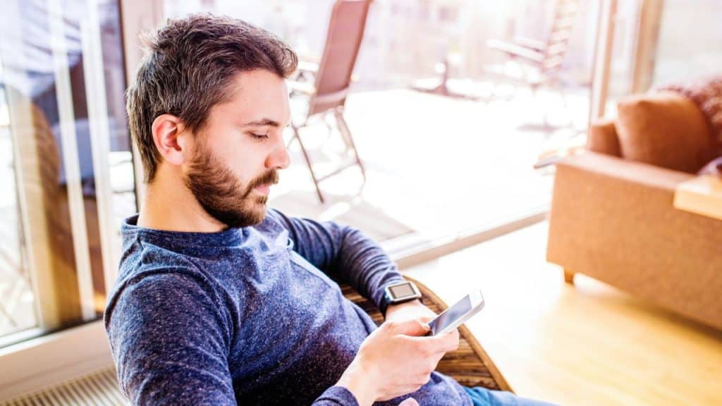 A man with a beard sits in a chair indoors and looks at his smartphone.