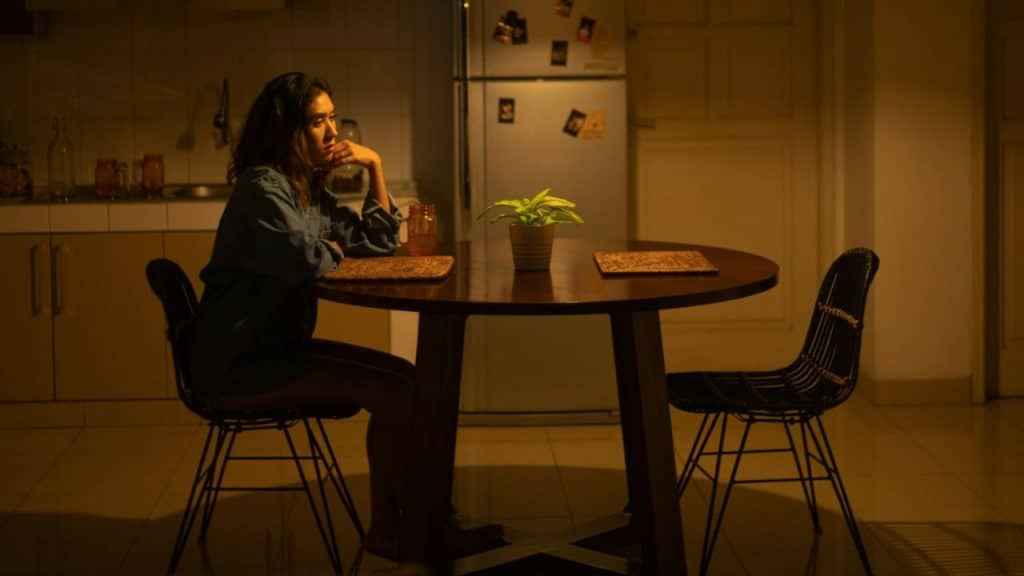 A woman sits alone at a dimly lit dining table in a kitchen at night.