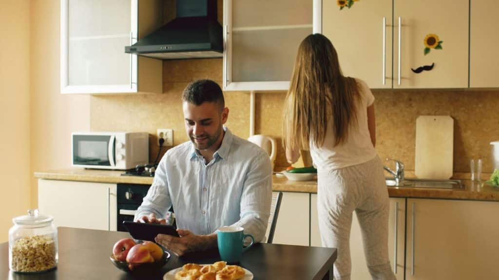 A man at a table uses a tablet while a woman works in the kitchen background.