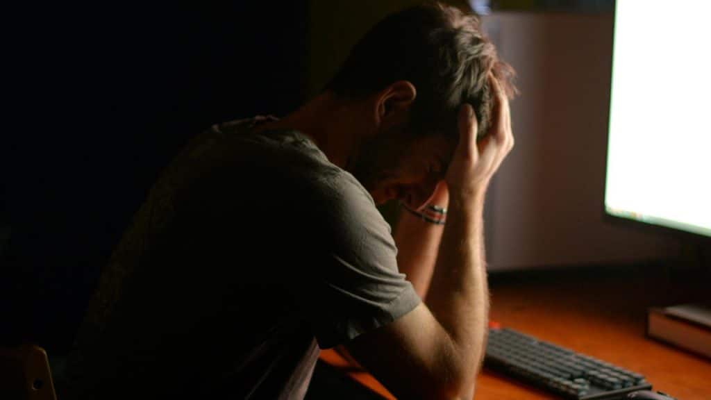A person sitting at a desk with their head in one hand, lit by a computer screen in a dark room.