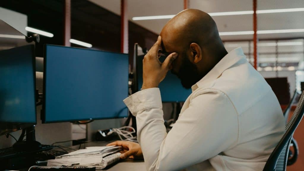 A bald man with a beard sits at a desk with computer monitors, touching his forehead.
