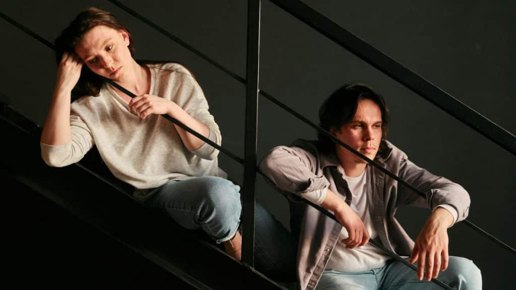 A man and woman sit on dark stairs behind metal railings looking in opposite directions.