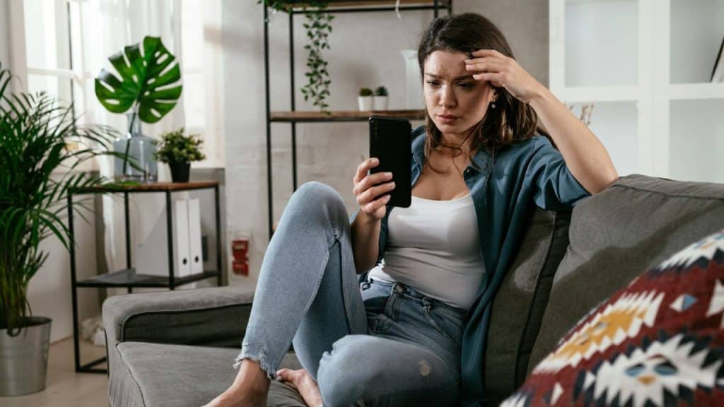 A woman sitting on a couch holds a phone and rests her hand on her forehead.