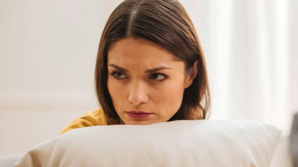 A woman in a yellow shirt rests her chin on a white pillow with a neutral expression.
