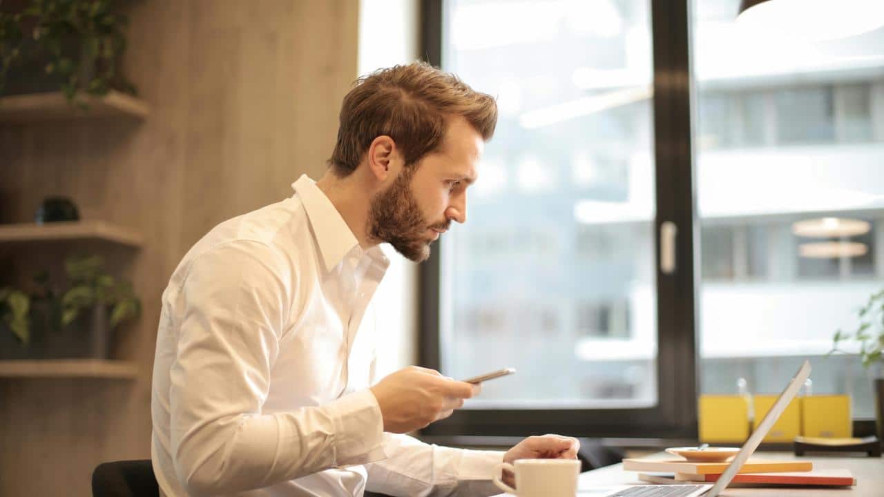 A man checking his phone at a laptop.