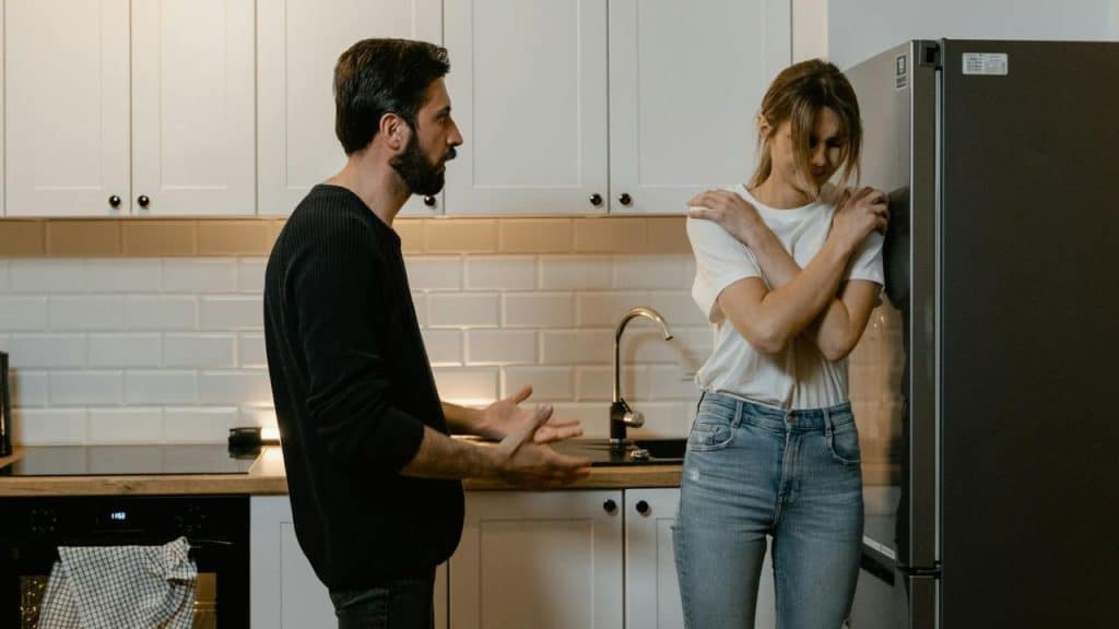 A bearded man gestures with both hands toward a woman with her arms crossed tightly.