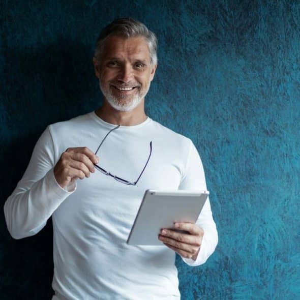 A smiling middle-aged man with gray hair holds a tablet and glasses against blue walls.