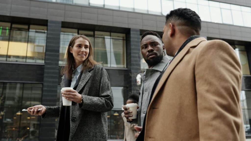 Three people in coats walk outside a modern building while holding paper coffee cups.