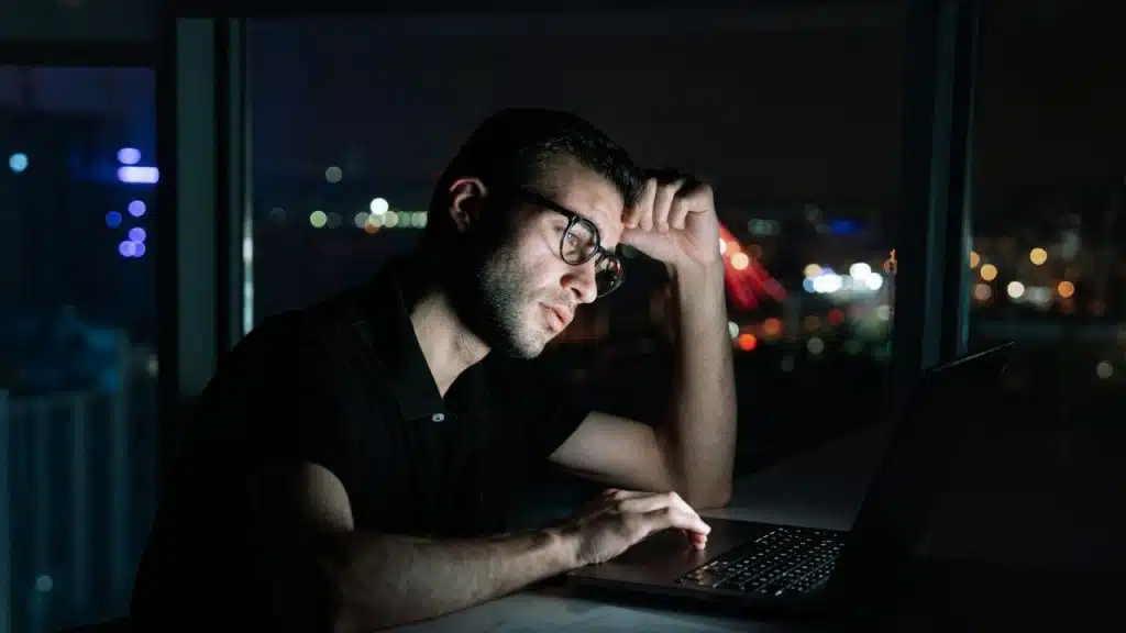 A man wearing glasses uses a laptop at night in front of a city window.