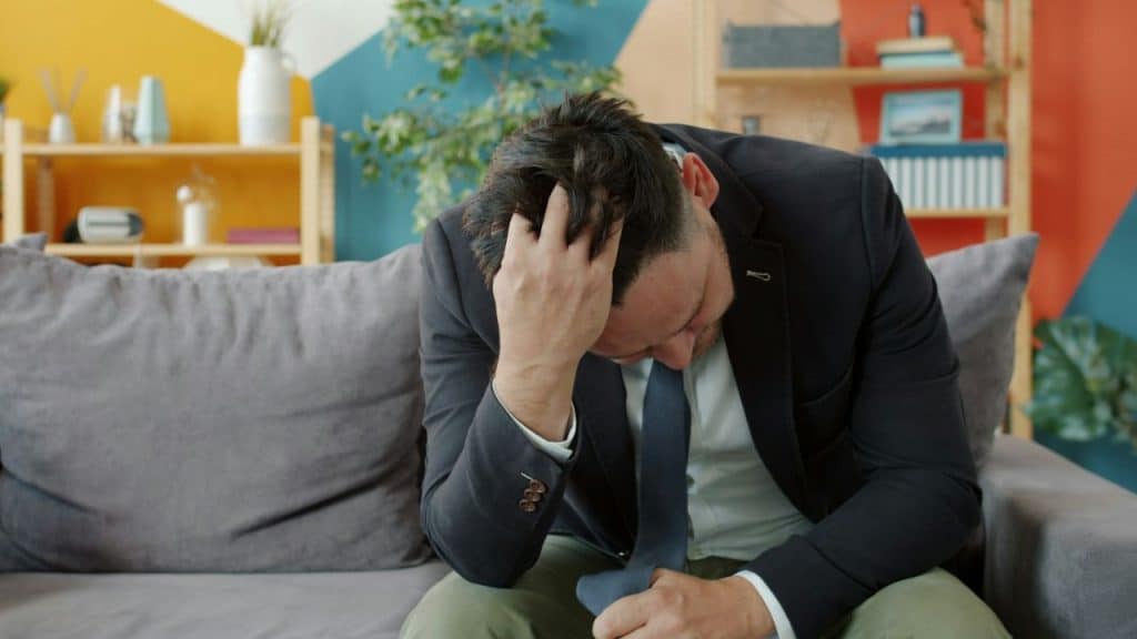 A man in a suit sits on a gray couch with his head down and hand in hair.