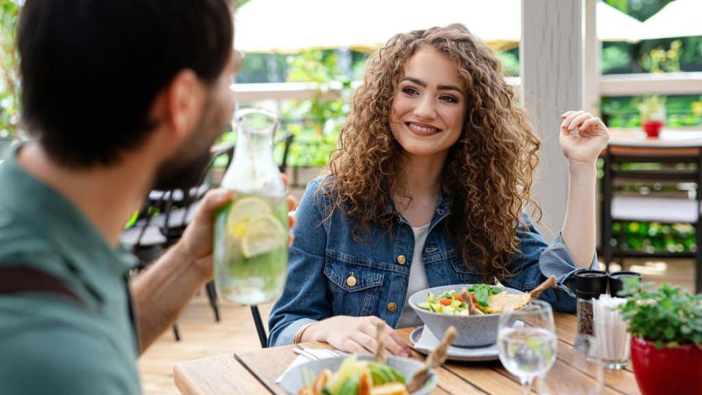 A woman smiling at a companion while enjoying a meal at an outdoor café.