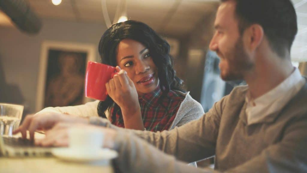 A woman holding a mug and talking with a man at a desk.
