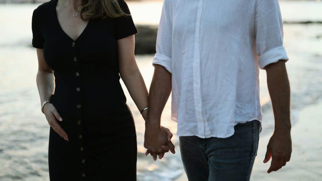A man and woman hold hands while walking along a beach during a sunset.