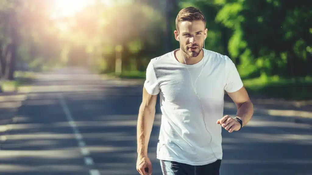 A man wearing a white t-shirt and earphones runs outdoors on a paved road.