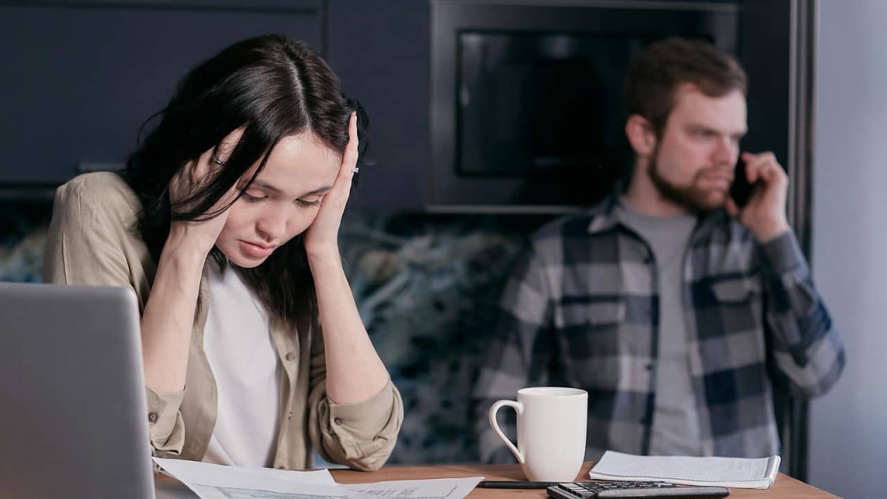A woman holds her head while looking at papers as a man speaks on a phone.