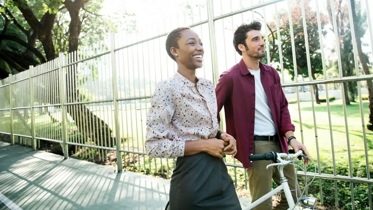 A man and woman walk alongside a bicycle past a metal fence in a park.