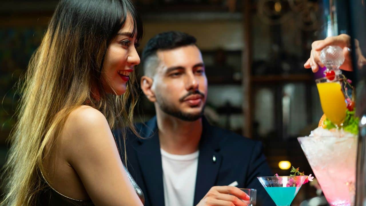 A woman smiles while sitting at a bar with a man as a drink is prepared.