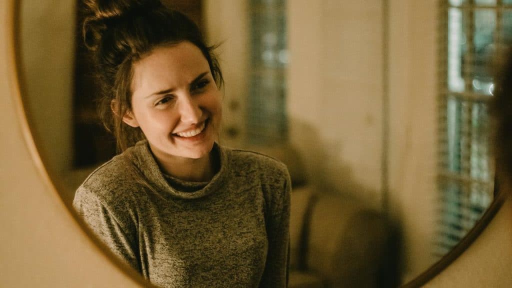 A smiling woman looking at her reflection in a softly lit room.