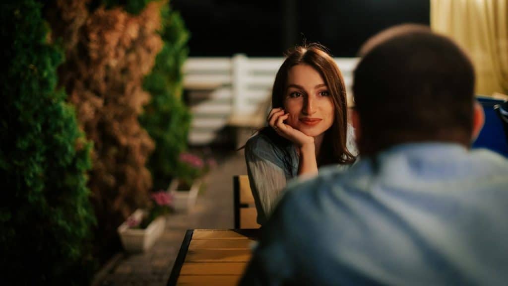 A woman sits at an outdoor table at night, resting her chin on her hand.