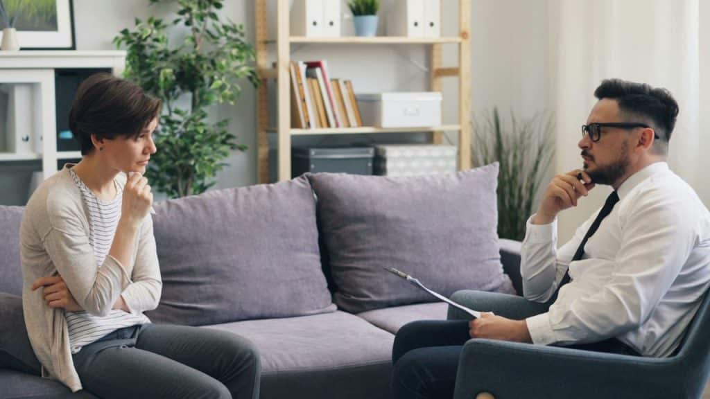 A woman sits on a sofa across from a man holding a clipboard and a pen.