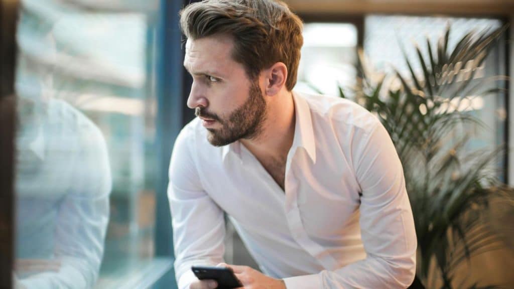 A man in a white shirt looks out a window while holding a black smartphone.