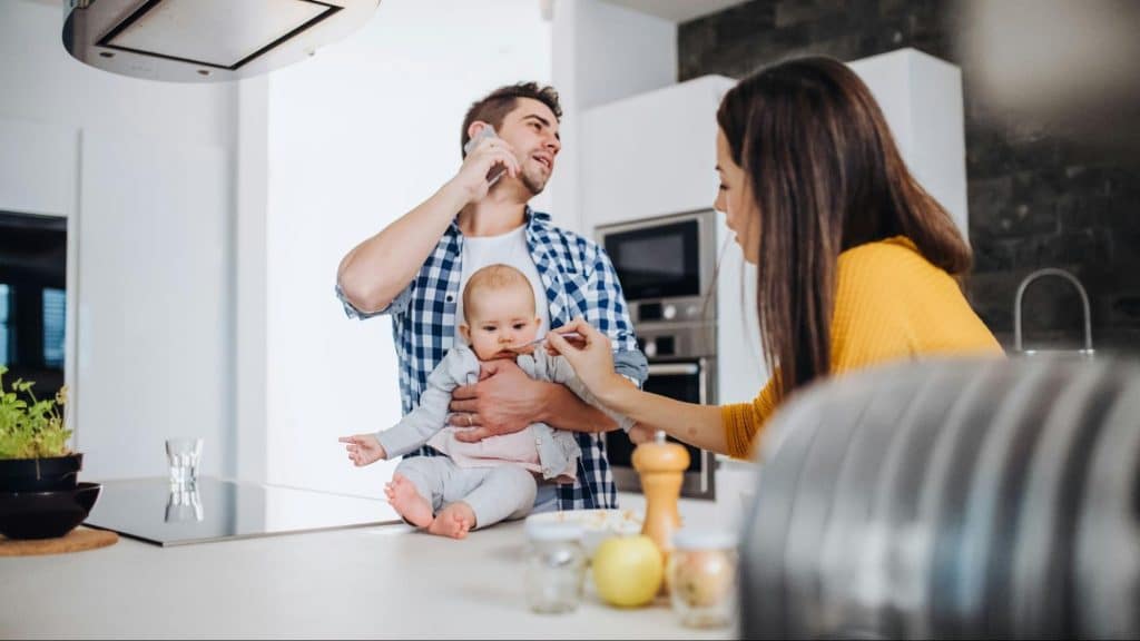 A man with smartphone and holding a baby while a woman feeds the baby