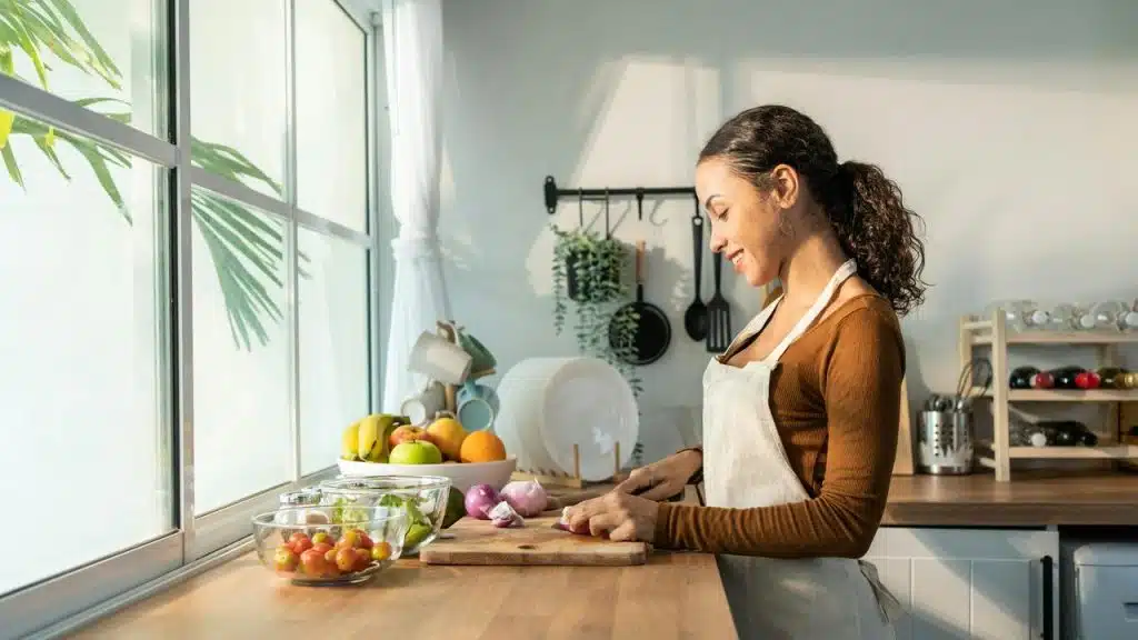 A woman in an apron chopping vegetables in a bright kitchen.