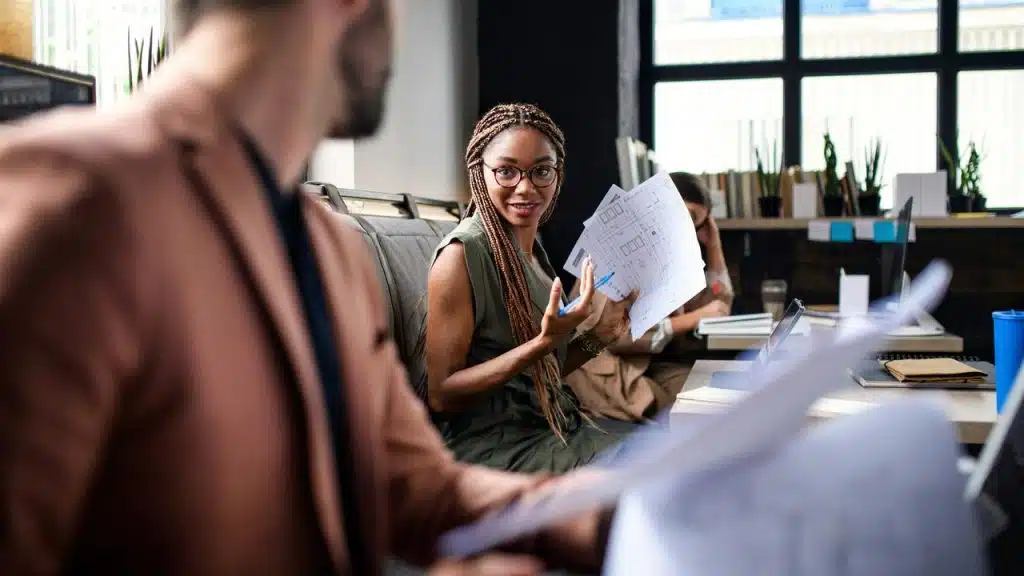 A woman holding papers and talking to a colleague in an office.
