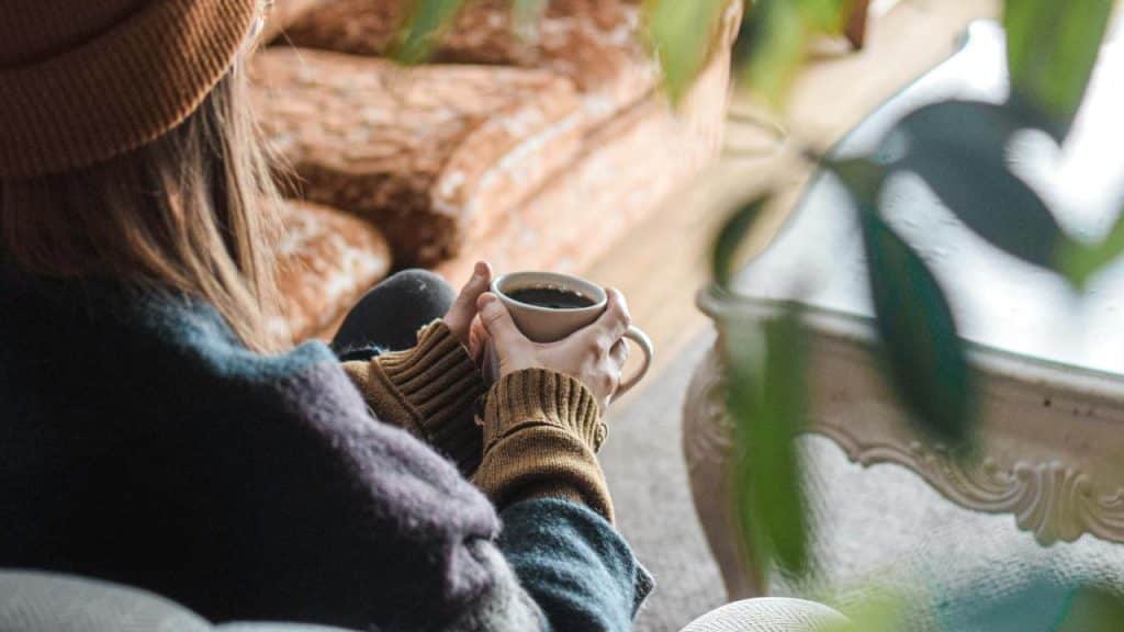 A person wearing a beanie and sweater holding a cup of coffee indoors.