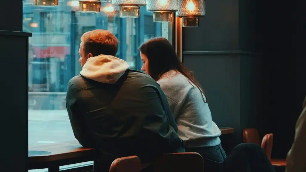 Two people sit side-by-side at a window counter inside a cafe looking outward.