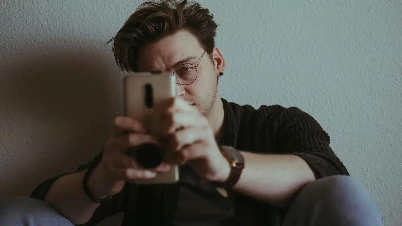 A man wearing glasses sits against a wall while holding a smartphone with both hands.