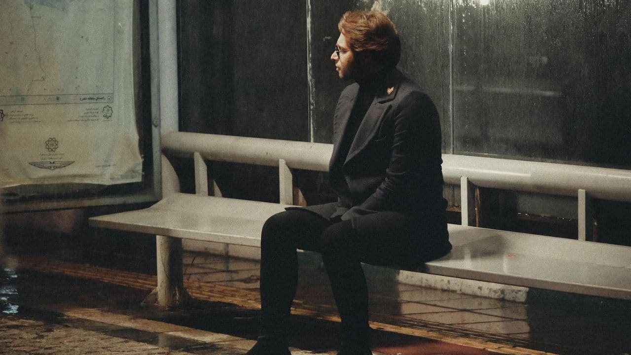 A man in dark clothing and glasses sits alone on a bench in the dark.