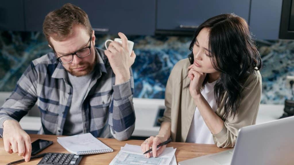 A man and woman sit at a desk covered in papers, a calculator, and a laptop.