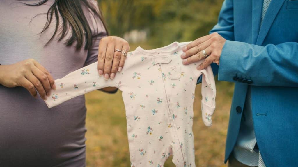 Two people stand outdoors holding a small floral infant onesie between them at chest height.