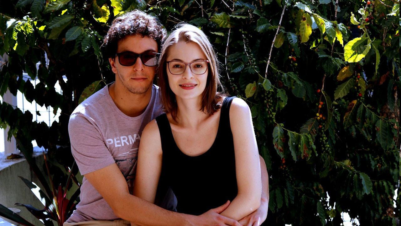 A man wearing sunglasses holding a woman wearing glasses closely as they sit in front of a leafy bush.