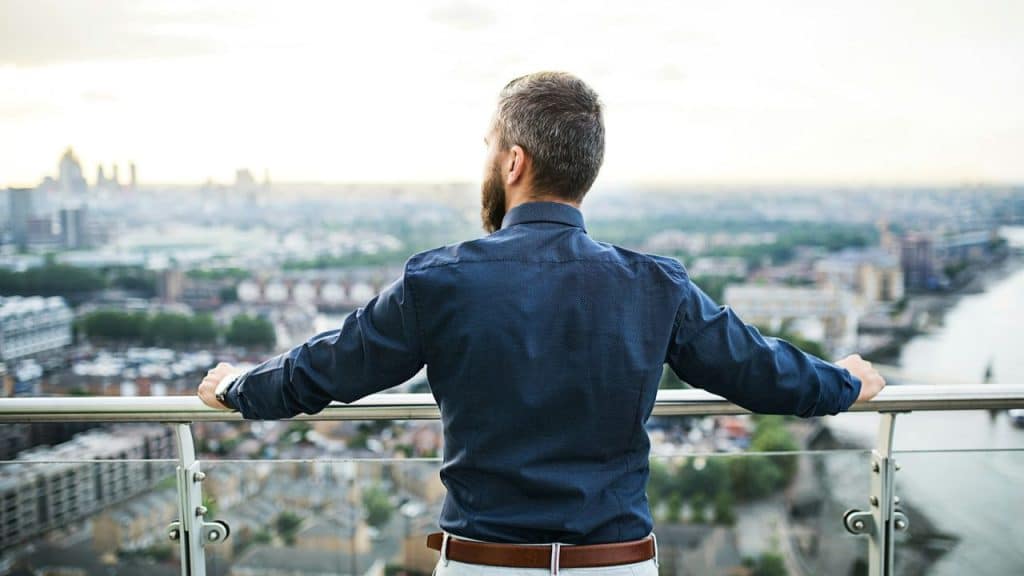 A man in a blue button-down shirt looks out over a city from a high balcony.