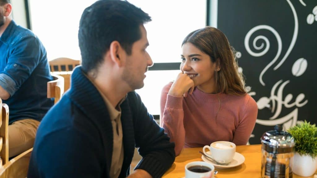 A woman in a pink sweater and a man in a navy cardigan talk over coffee.