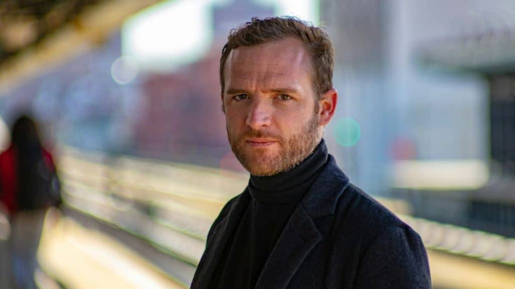 A man in a black turtleneck and dark jacket looks directly at the camera outdoors.