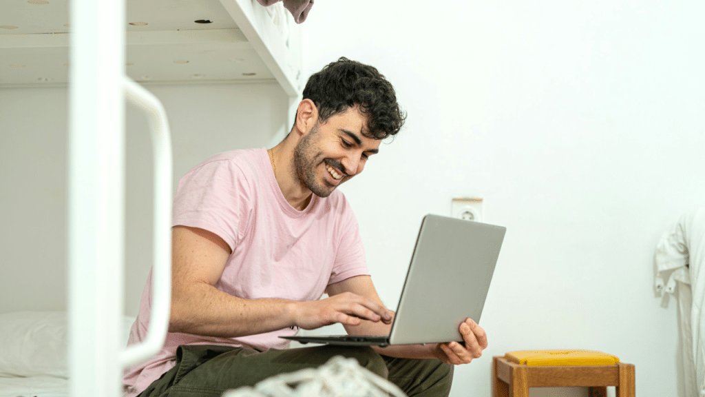 A smiling man in a pink t-shirt sits on a bed while using a laptop computer.