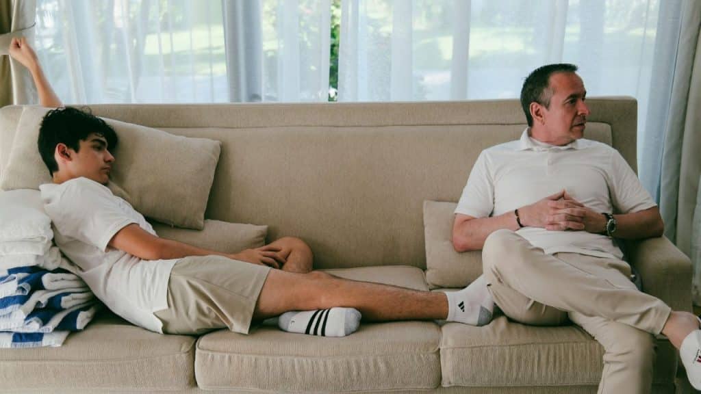 A man and a teenager sit on opposite ends of a long couch looking away from each other.